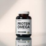 A sleek and modern protein supplement bottle, showcasing the "Protein Omega" label in a clean, minimalist design. The bottle is positioned on a reflective, glass-like surface, casting a subtle shadow below. Soft, directional lighting illuminates the supplement from the side, creating a sense of depth and dimension. The overall scene has a professional, high-quality aesthetic, inviting the viewer to imagine the premium quality of the product. The background is a simple, neutral color that allows the supplement bottle to be the focal point, drawing the eye to the key details and branding.