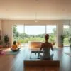 Peaceful wellness services center set in a serene natural environment. Sunlit interior with clean, minimalist decor - wooden floors, neutral tones, plants, and soft lighting. In the foreground, individuals engage in yoga, meditation, and other holistic practices. In the middle ground, a reception area with a calming water feature. The background features lush greenery, a tranquil pond, and a clear sky with wispy clouds. An atmosphere of tranquility, balance, and rejuvenation permeates the scene.